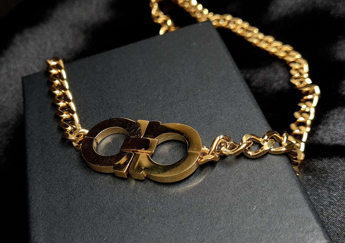 A gold chain necklace with the letter C on it sitting on top of a black box. The necklace is made of gold and has a simple design. The letter C is large and has a polished finish. The necklace is elegant and understated.