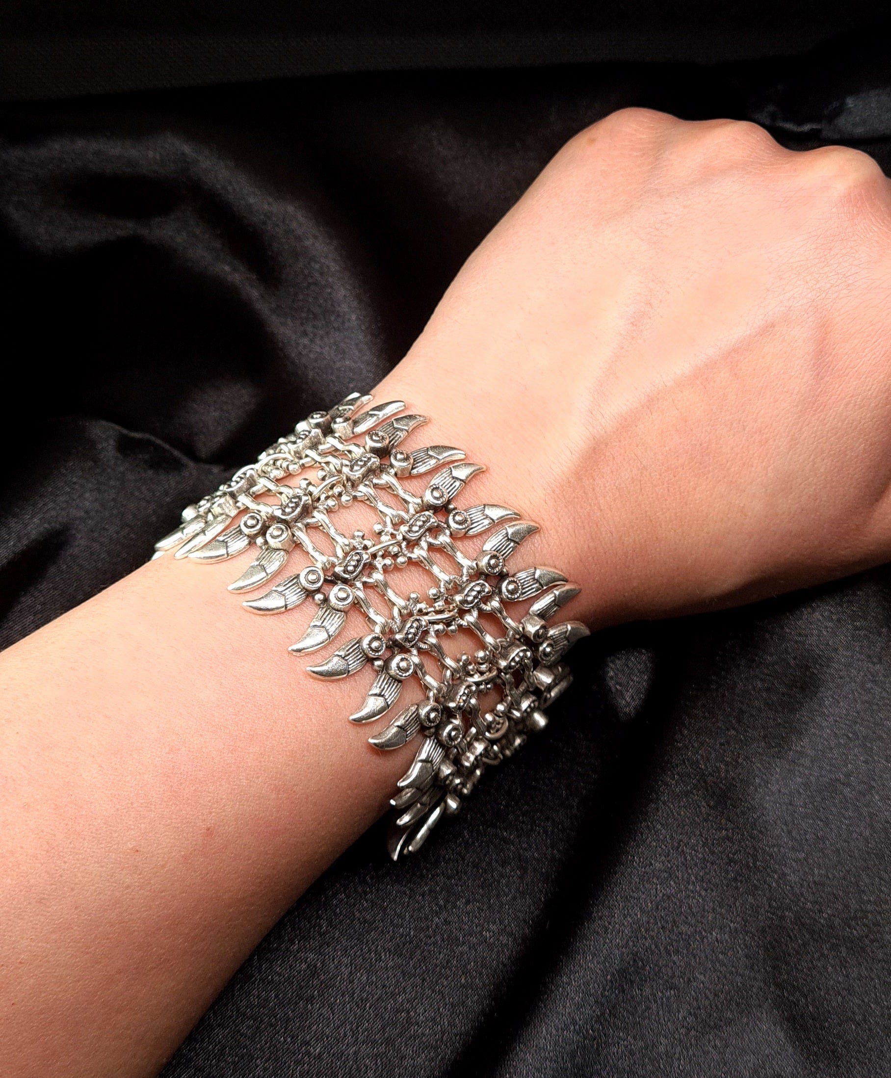 a woman hand wearing A silver bracelet with a claw design on it. The bracelet is made of silver and has a delicate design. The claw design is in the center of the bracelet and is made up of small, interlocking pieces. The bracelet is sitting on a black cloth