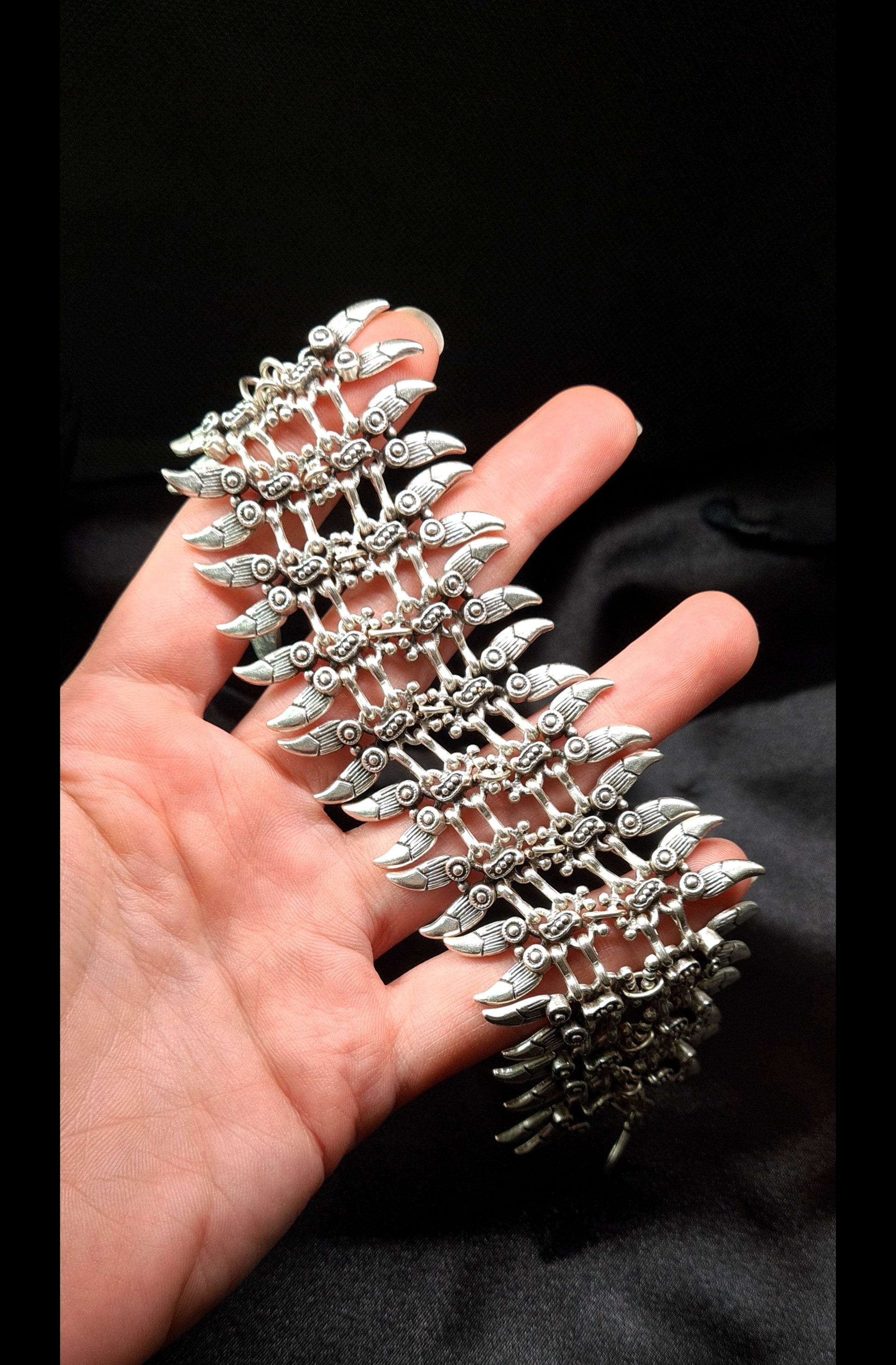 a hand holding A silver bracelet with a claw design on it. The bracelet is made of silver and has a delicate design. The claw design is in the center of the bracelet and is made up of small, interlocking pieces. The bracelet is sitting on a black cloth