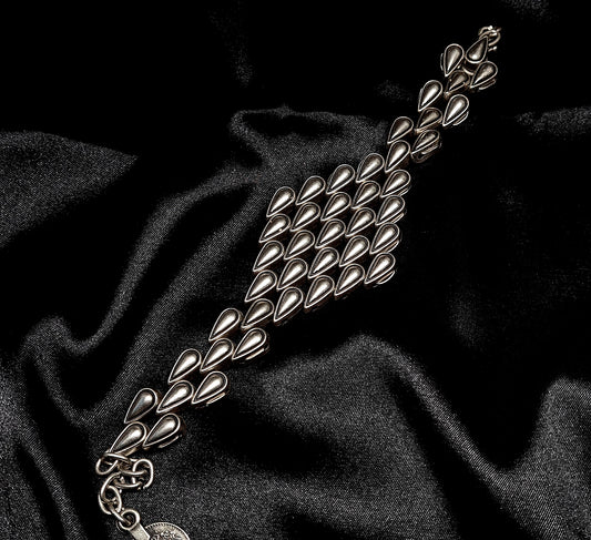 A silver bracelet with a tear drops shaped pendant on it. The bracelet is made of silver and has a delicate design. The shaped pendant is small and has a smooth finish. The bracelet is sitting on a black cloth.