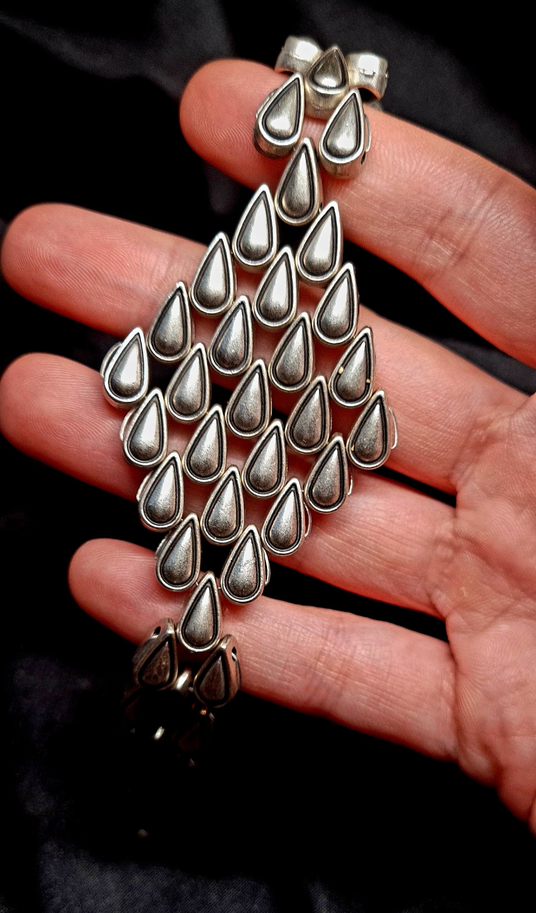 a hand holding A silver bracelet with a tear drops shaped pendant on it. The bracelet is made of silver and has a delicate design. The shaped pendant is small and has a smooth finish. The bracelet is sitting on a black cloth.