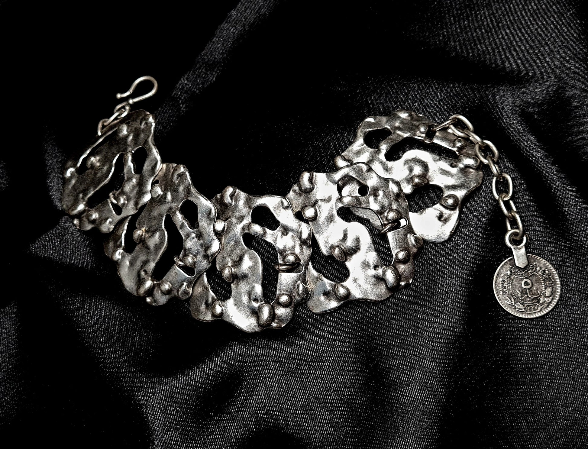 A silver bracelet sitting on a black cloth. The bracelet is made of silver and has a delicate design. The coin is small and round, and it has a silver color. The bracelet is on a black cloth.