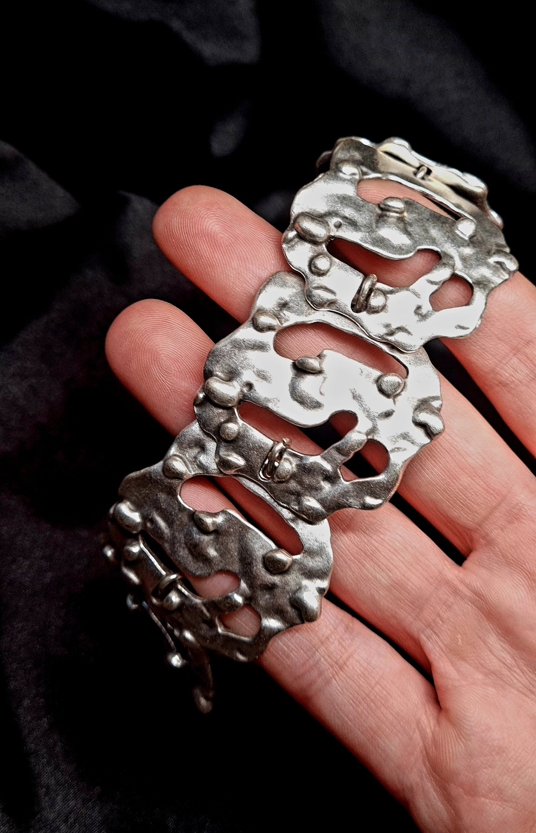 a hand holding A silver bracelet sitting on a black cloth. The bracelet is made of silver and has a delicate design. The coin is small and round, and it has a silver color. The bracelet is on a black cloth.