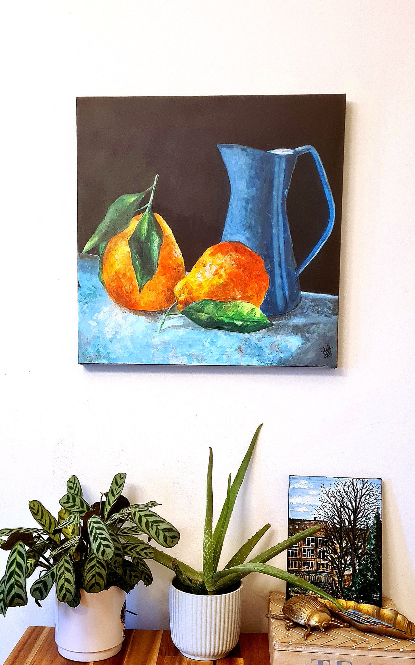 A painting of pears and a blue pitcher hanging on a wall next to potted plants on a table. The painting is done in a realistic style, with attention to detail. The pears are a vibrant orange color. The pitcher is a blue color, and the table is a wooden table. The potted plants are green. The painting is hung on a white wall