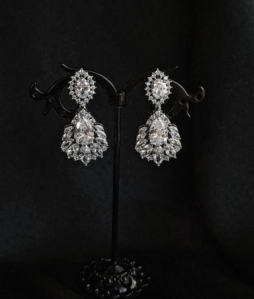 A pair of earrings with diamonds on a black background. The earrings are made of silver and have a delicate design. The diamonds are round and sparkling. The earrings are hanging from a silver chain.