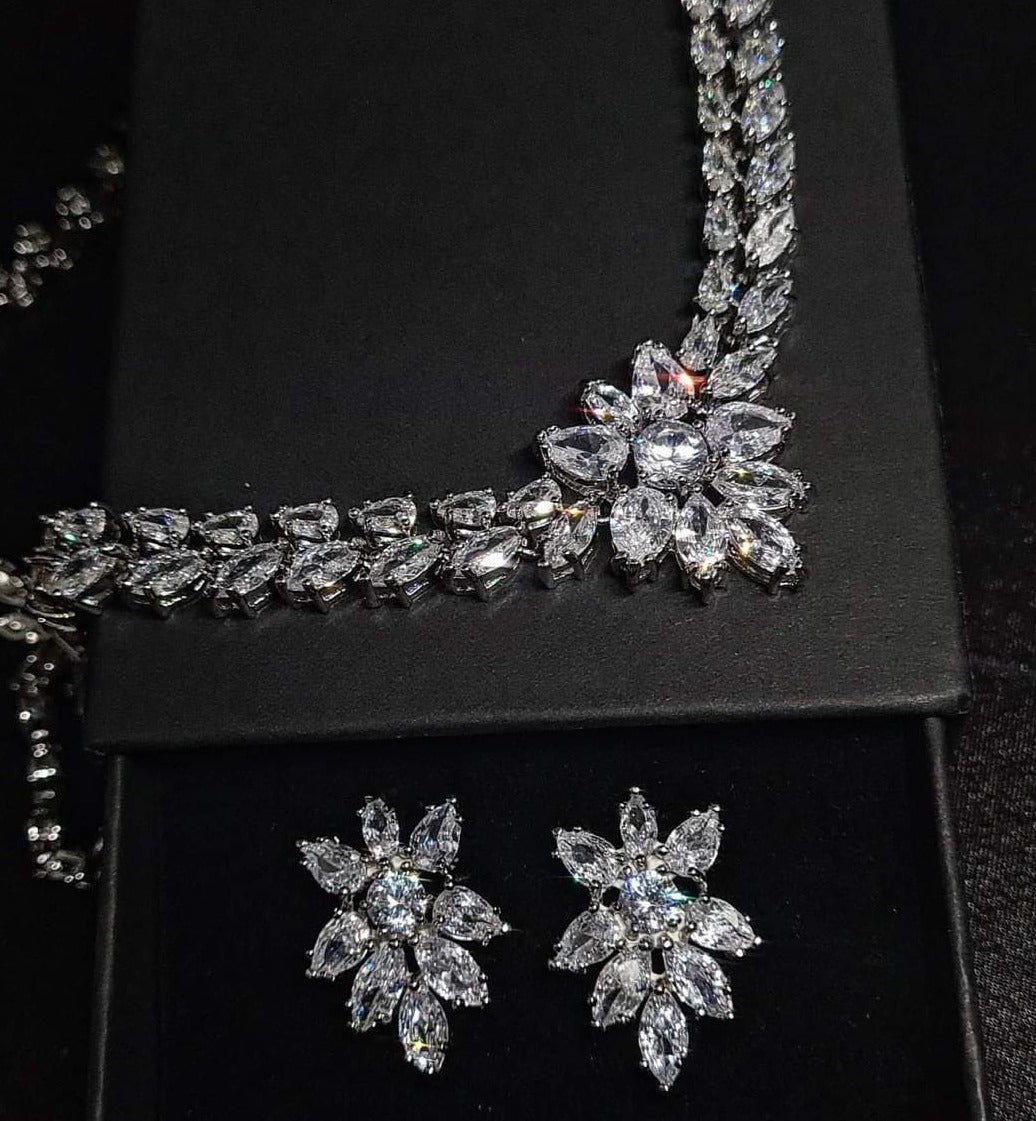Wedding necklace and earrings set on a mannequin. The necklace is a long, flowing piece with a delicate design. It is made of silver with sparkling crystals. The earrings are matching flower shapes with a similar design, The necklace and earrings are displayed on a black mannequin. The background is also black.