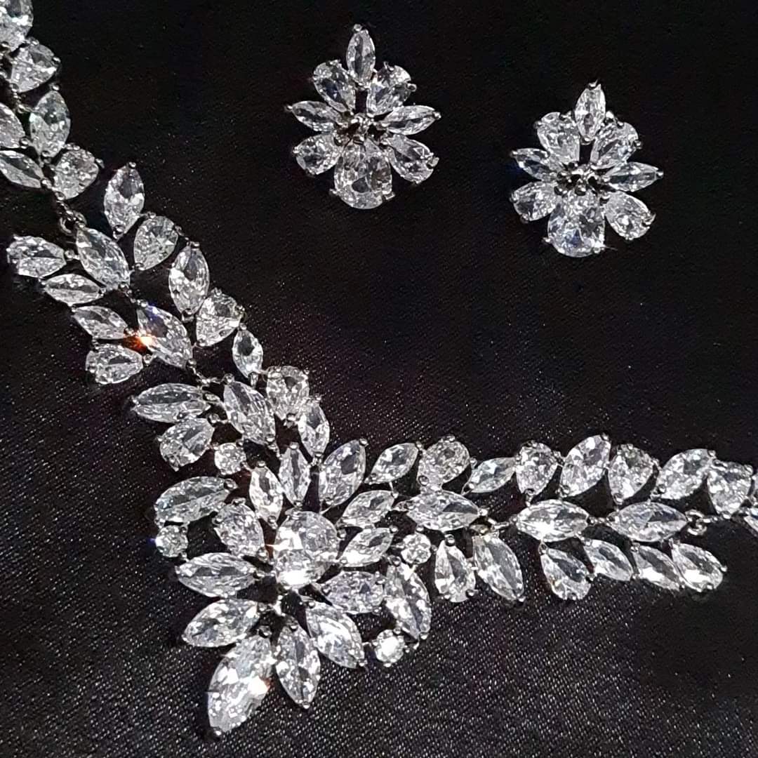 Wedding necklace and earrings set on a mannequin. The necklace is a long, flowing piece with a delicate design. It is made of silver with sparkling crystals. The earrings are matching teardrop shapes with a similar design, The necklace and earrings are displayed on a black mannequin. The background is also black.