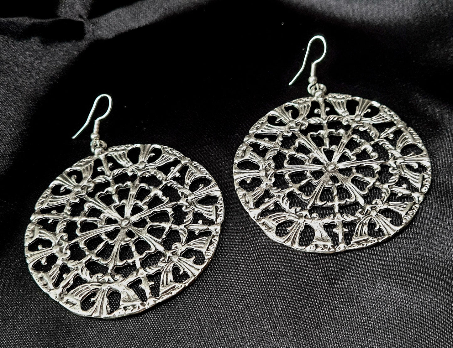 A pair of silver earrings on a black surface. The earrings are made of silver and have a circular design on them. The earrings are small and delicate, and they have a shiny finish.