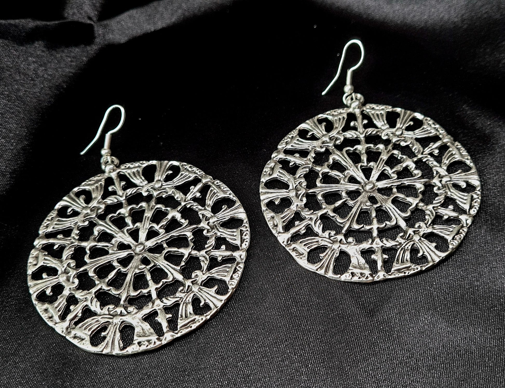 A pair of silver earrings on a black surface. The earrings are made of silver and have a circular design on them. The earrings are small and delicate, and they have a shiny finish.