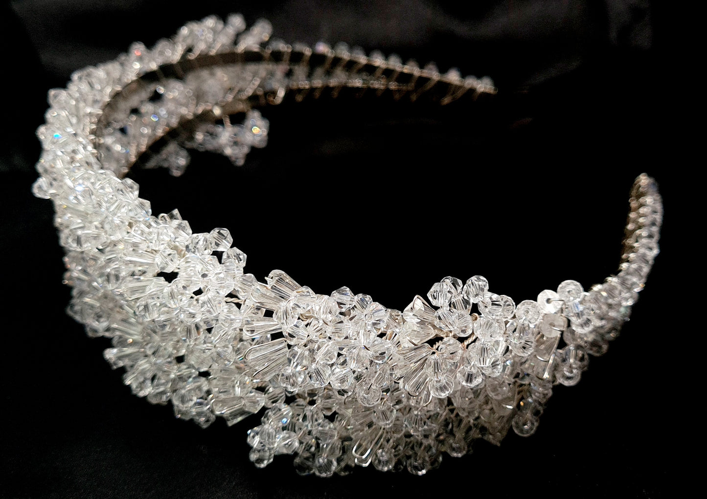 A close-up of a headband with crystals on a black background. The headband is made of a silver-colored metal and has an intricate design with crystals of different shapes and sizes. The crystals are sparkling and reflect the light.