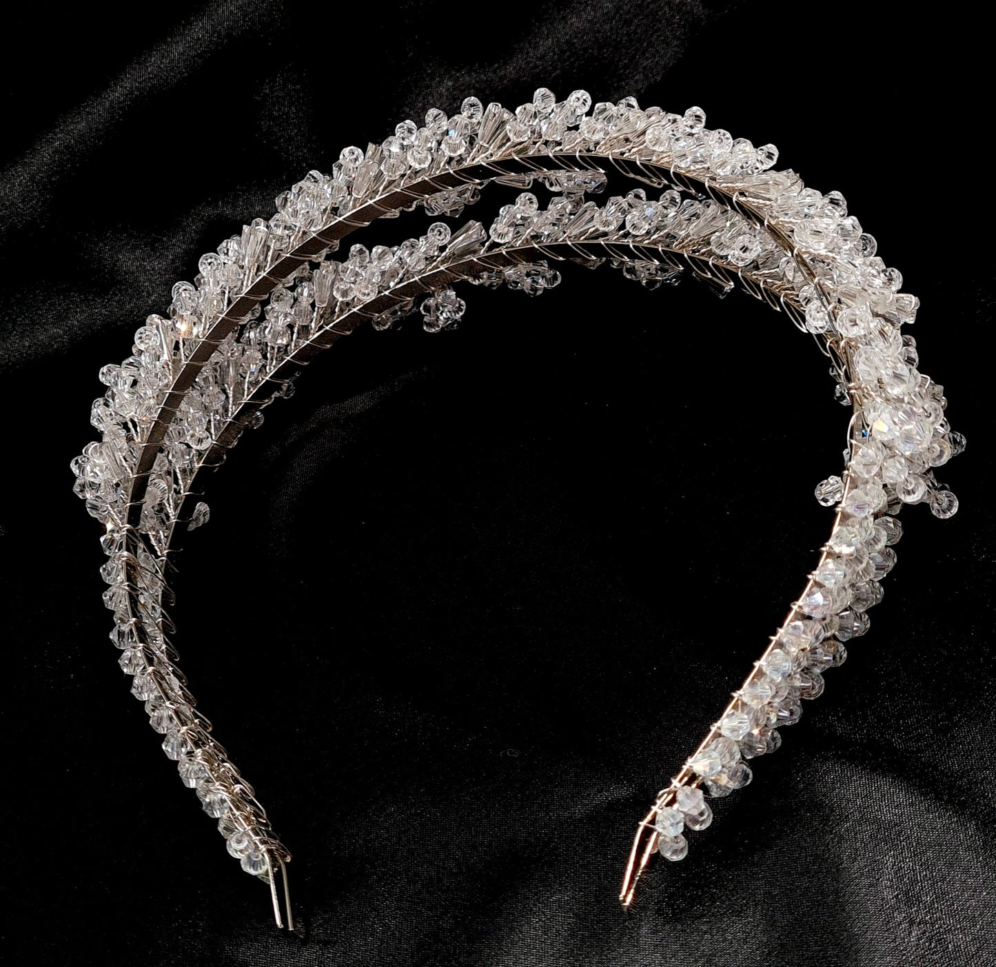 A close-up of a headband with crystals on a black background. The headband is made of a silver-colored metal and has an intricate design with crystals of different shapes and sizes. The crystals are sparkling and reflect the light.