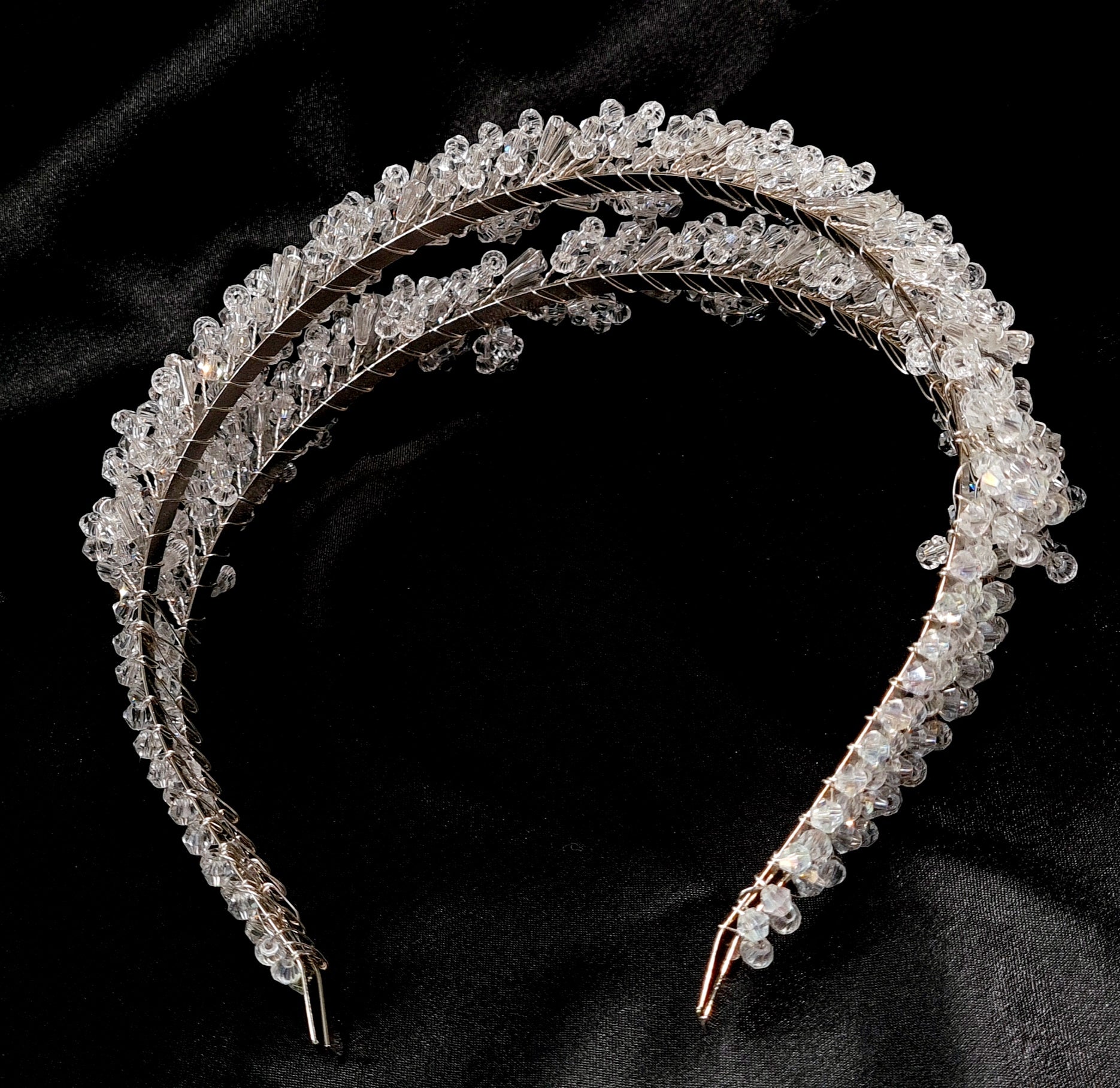 A close-up of a headband with crystals on a black background. The headband is made of a silver-colored metal and has an intricate design with crystals of different shapes and sizes. The crystals are sparkling and reflect the light.
