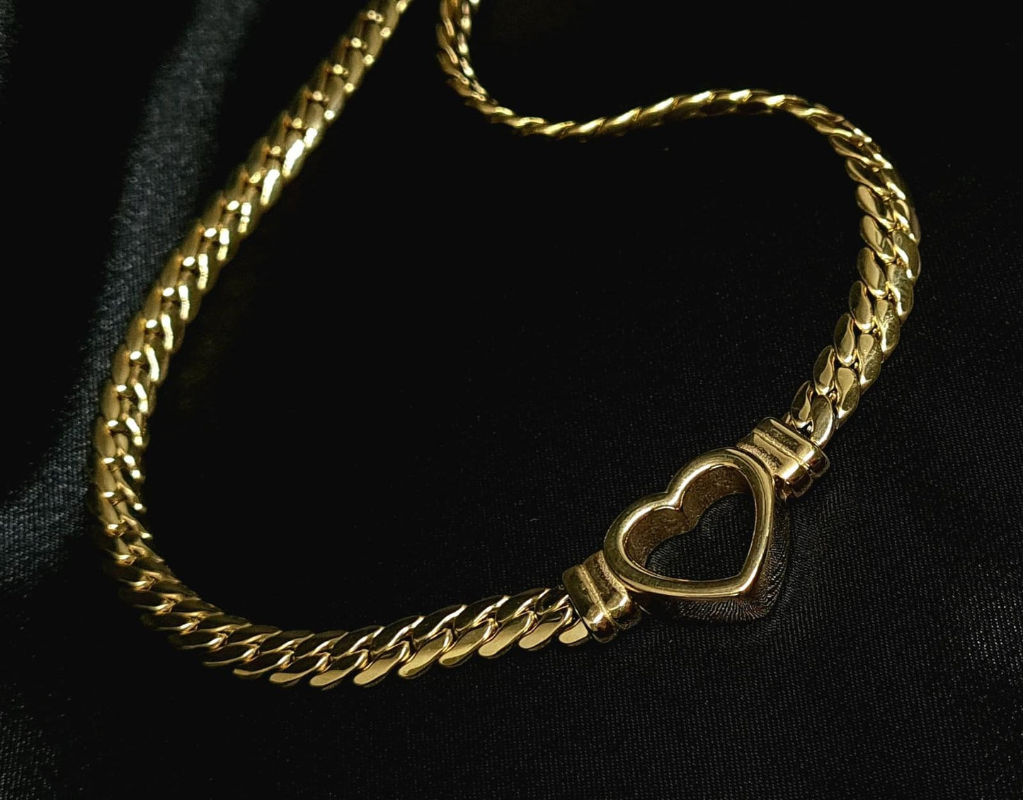 Gold necklace with heart pendant on black background. The necklace is made of gold and has a heart pendant in the center. The heart is gold and has a smooth surface. The necklace is sitting on a black background, which provides a nice contrast to the gold color of the heart