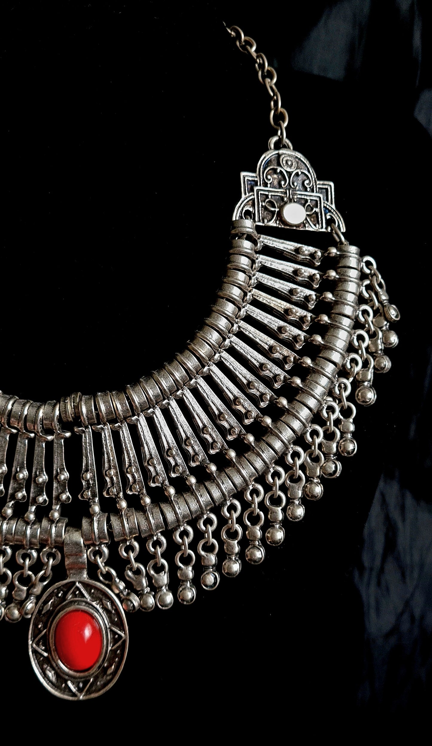close up view of A silver and red necklace on a black background. The necklace is a long, filigree chain with a drop-shaped pendant. The pendant is made of silver and has a red stone in the center. The stone is a deep red color and has a polished finish. The background is a black velvet cloth.