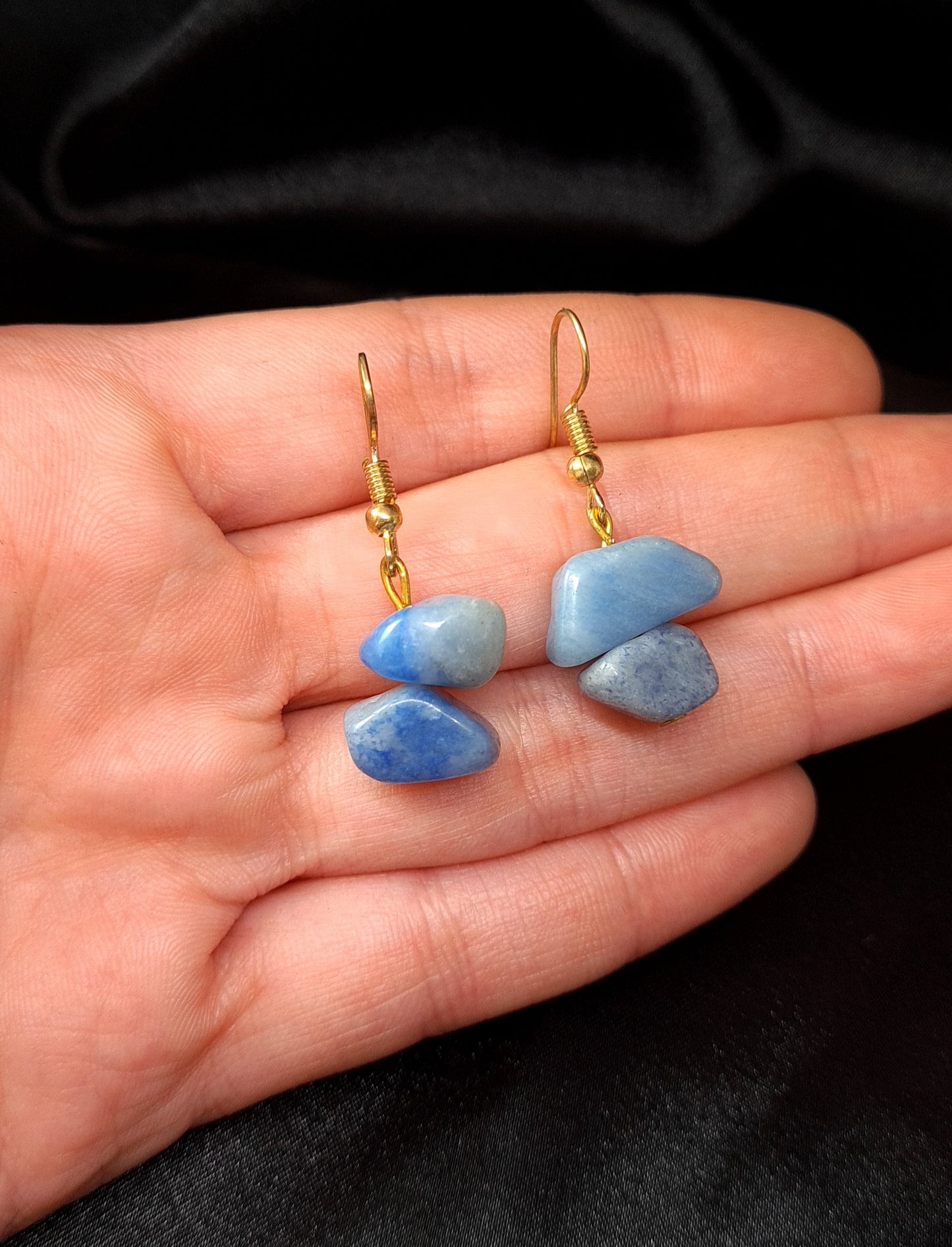 a hand holding Pair of blue earrings with gold hooks. The earrings are sitting on a black box. The earrings are made of blue rocks and gold hooks. The rocks are oval-shaped and have a smooth surface. The earrings are a beautiful and stylish accessory that would be perfect for any occasion.