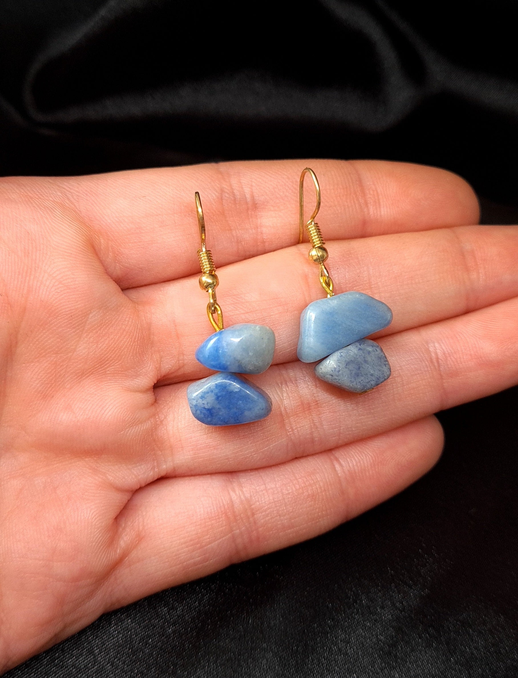a hand holding Pair of blue earrings with gold hooks. The earrings are sitting on a black box. The earrings are made of blue rocks and gold hooks. The rocks are oval-shaped and have a smooth surface. The earrings are a beautiful and stylish accessory that would be perfect for any occasion.