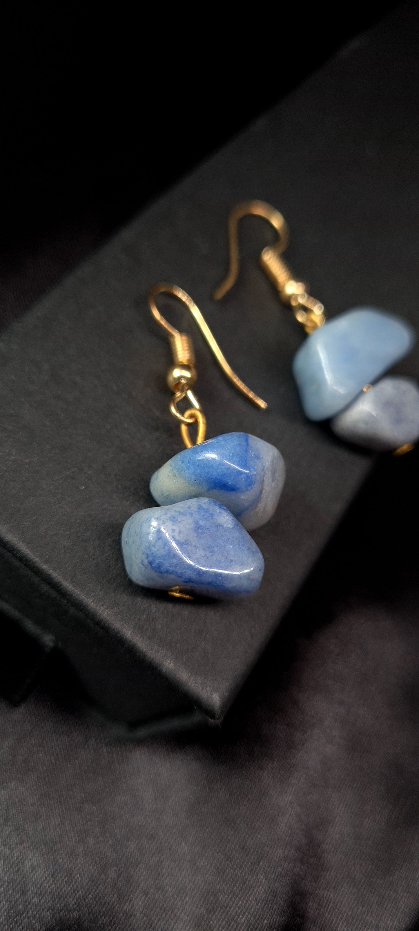 Pair of blue earrings with gold hooks. The earrings are sitting on a black box. The earrings are made of blue rocks and gold hooks. The rocks are oval-shaped and have a smooth surface. The earrings are a beautiful and stylish accessory that would be perfect for any occasion.