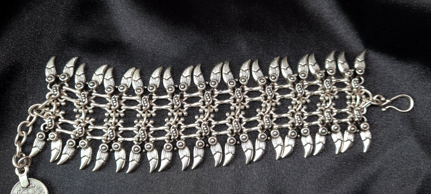A silver bracelet with a claw design on it. The bracelet is made of silver and has a delicate design. The claw design is in the center of the bracelet and is made up of small, interlocking pieces. The bracelet is sitting on a black cloth