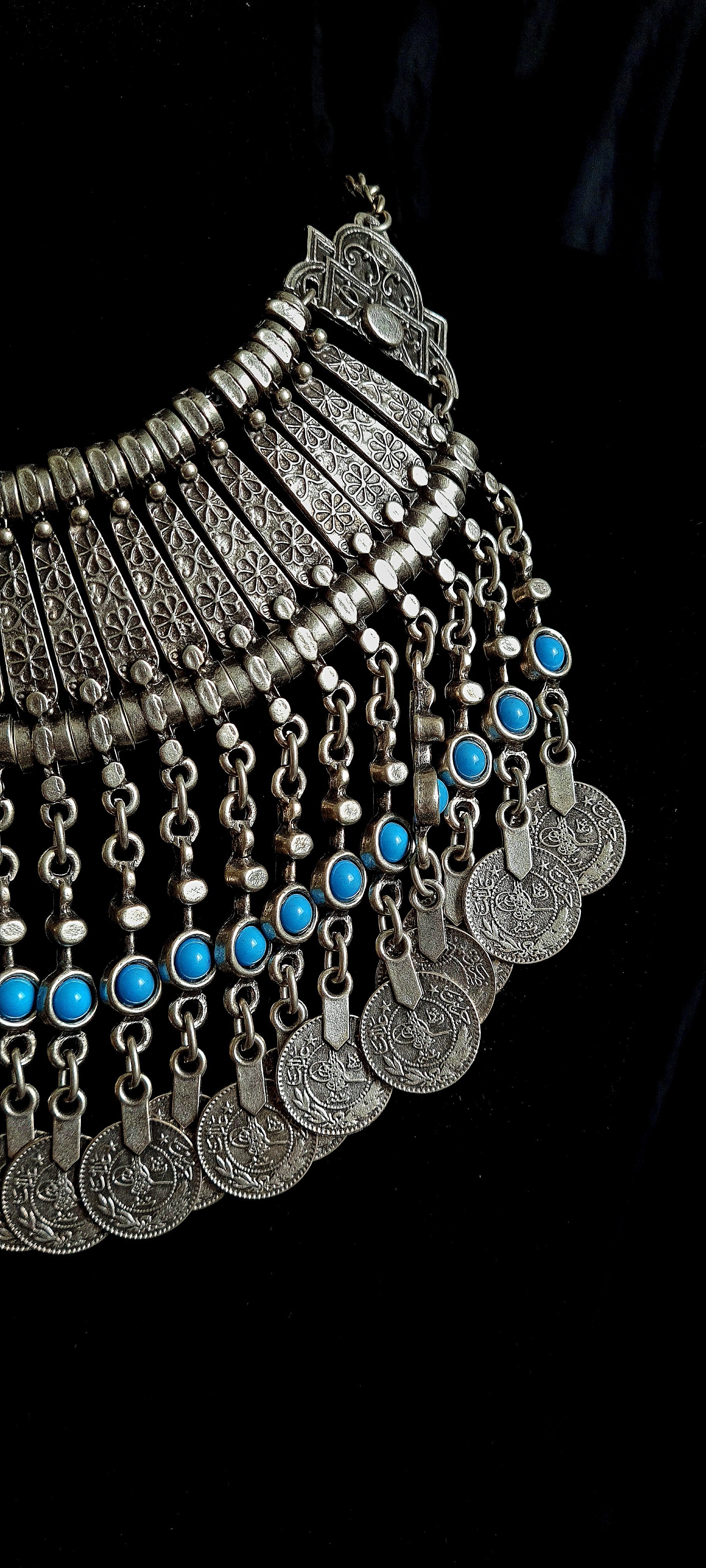 close up view of A silver necklace with turquoise beads and coins on a black background. The necklace is made of silver and has a delicate design. The turquoise beads are small and round, and they have a bright blue color. The coins are also small and round, and they have a bronze color. The necklace is long and hangs down to the chest.