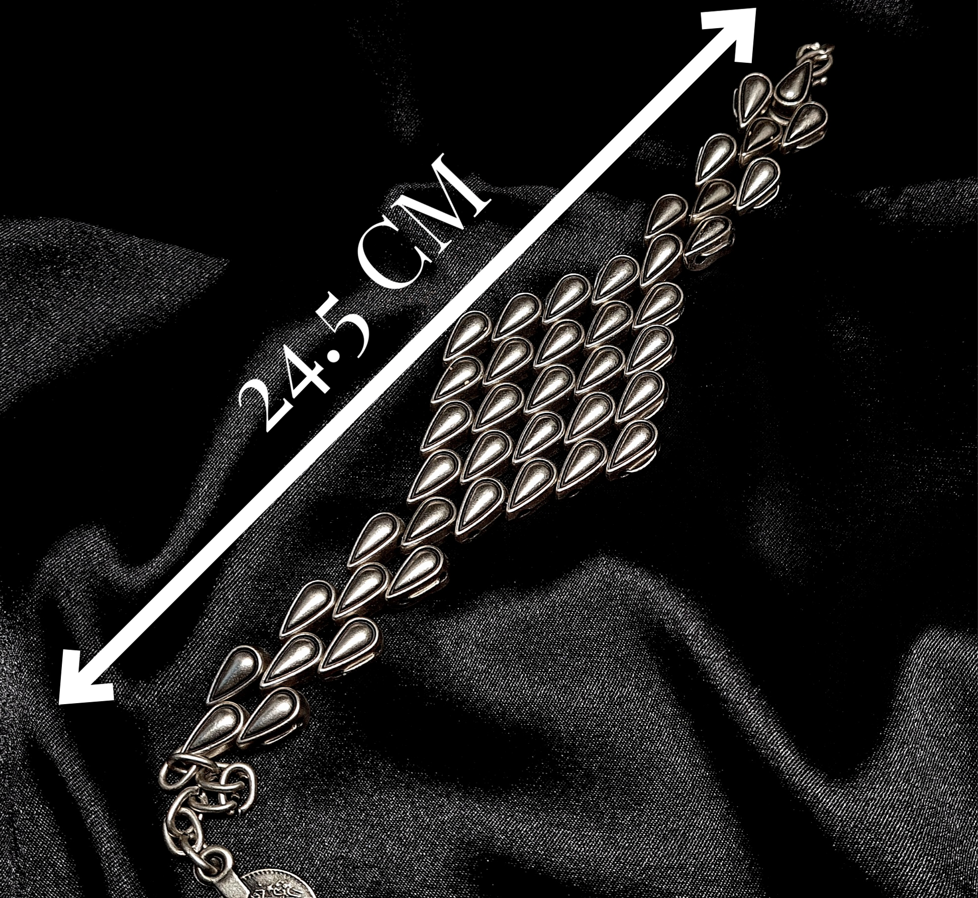 A silver bracelet with a tear drops shaped pendant on it. The bracelet is made of silver and has a delicate design. The shaped pendant is small and has a smooth finish. The bracelet is sitting on a black cloth with measures.