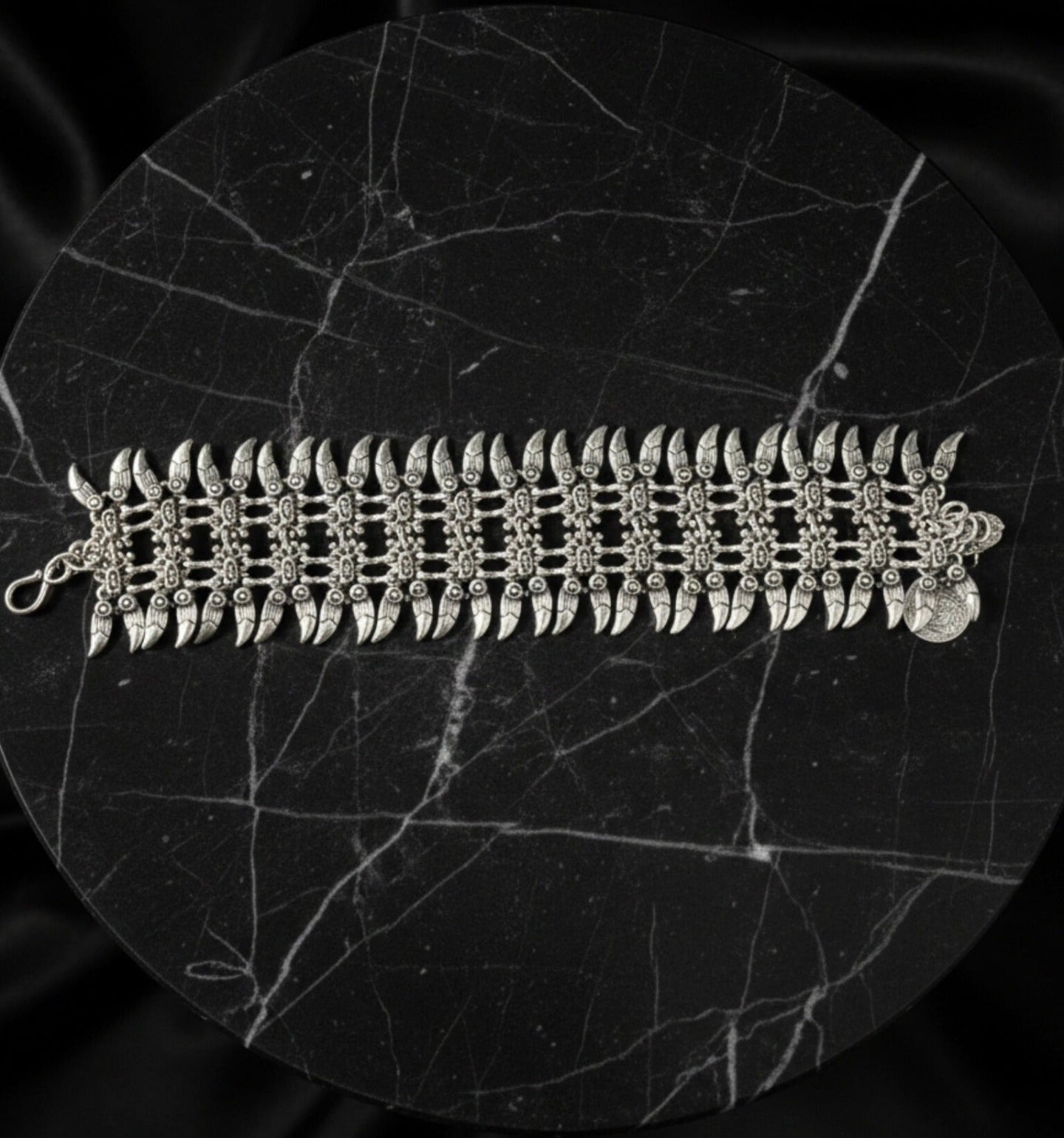 Luxury product display of a wide silver spike cuff bracelet laid flat on a circular black marble plinth, showcasing the intricate metalwork and bohemian-inspired charms.