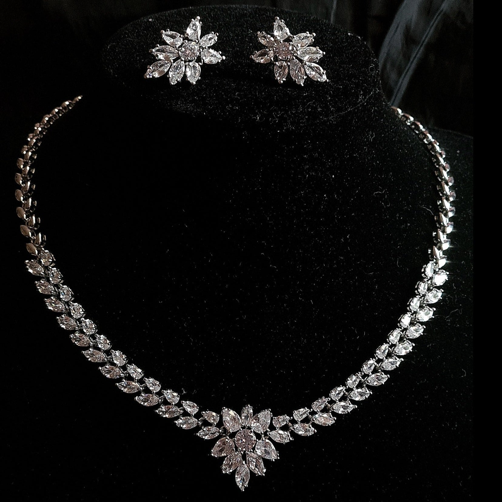 Wedding necklace and earrings set on a mannequin. The necklace is a long, flowing piece with a delicate design. It is made of silver with sparkling crystals. The earrings are matching flower shapes with a similar design, The necklace and earrings are displayed on a black mannequin. The background is also black.
