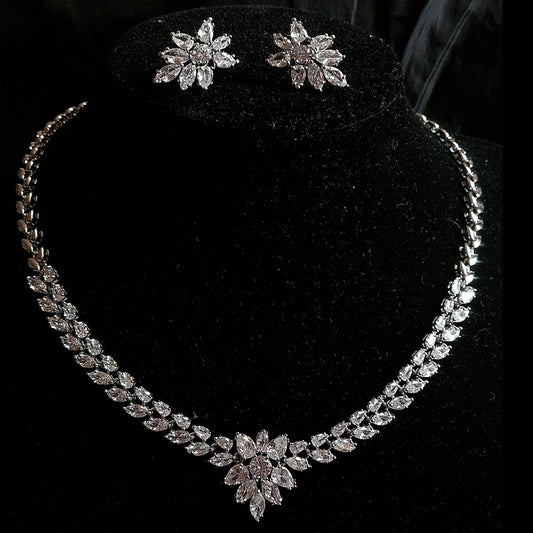 Wedding necklace and earrings set on a mannequin. The necklace is a long, flowing piece with a delicate design. It is made of silver with sparkling crystals. The earrings are matching flower shapes with a similar design, The necklace and earrings are displayed on a black mannequin. The background is also black.