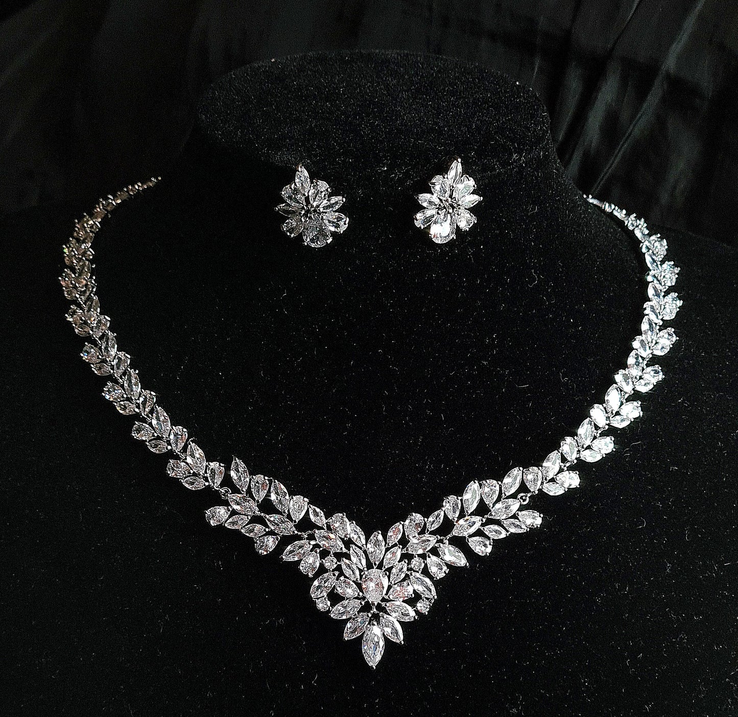 Wedding necklace and earrings set on a mannequin. The necklace is a long, flowing piece with a delicate design. It is made of silver with sparkling crystals. The earrings are matching teardrop shapes with a similar design, The necklace and earrings are displayed on a black mannequin. The background is also black.