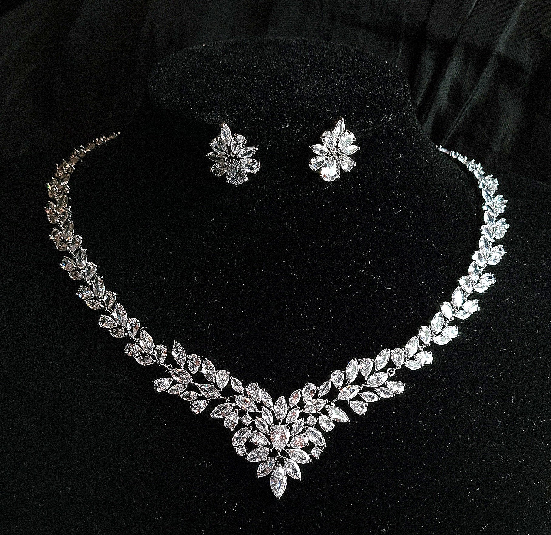 Wedding necklace and earrings set on a mannequin. The necklace is a long, flowing piece with a delicate design. It is made of silver with sparkling crystals. The earrings are matching teardrop shapes with a similar design, The necklace and earrings are displayed on a black mannequin. The background is also black.
