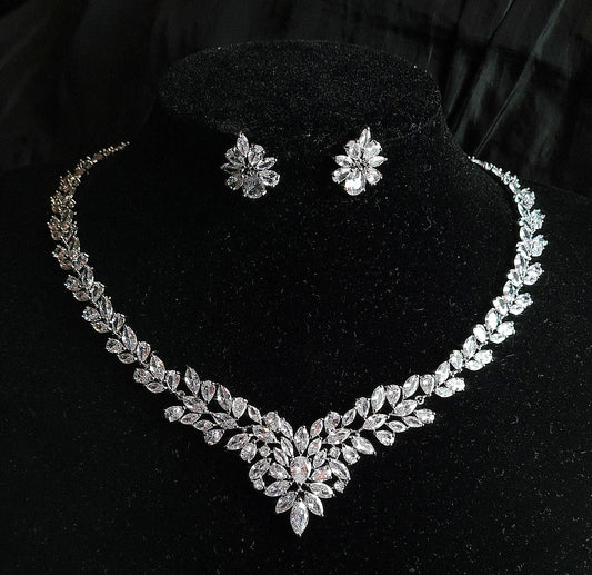 Wedding necklace and earrings set on a mannequin. The necklace is a long, flowing piece with a delicate design. It is made of silver with sparkling crystals. The earrings are matching teardrop shapes with a similar design, The necklace and earrings are displayed on a black mannequin. The background is also black.