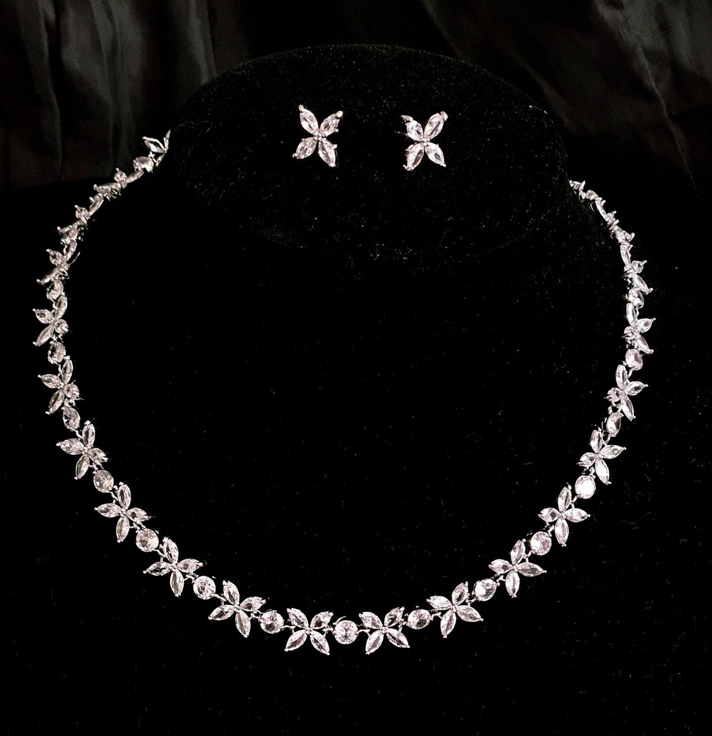 A silver necklace and earrings set with rhinestones on a black background. The necklace is long and has a delicate design. The earrings are small and have a flower shaped pendant. The rhinestones are sparkling and reflect the light.