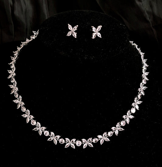 A silver necklace and earrings set with rhinestones on a black background. The necklace is long and has a delicate design. The earrings are small and have a flower shaped pendant. The rhinestones are sparkling and reflect the light.