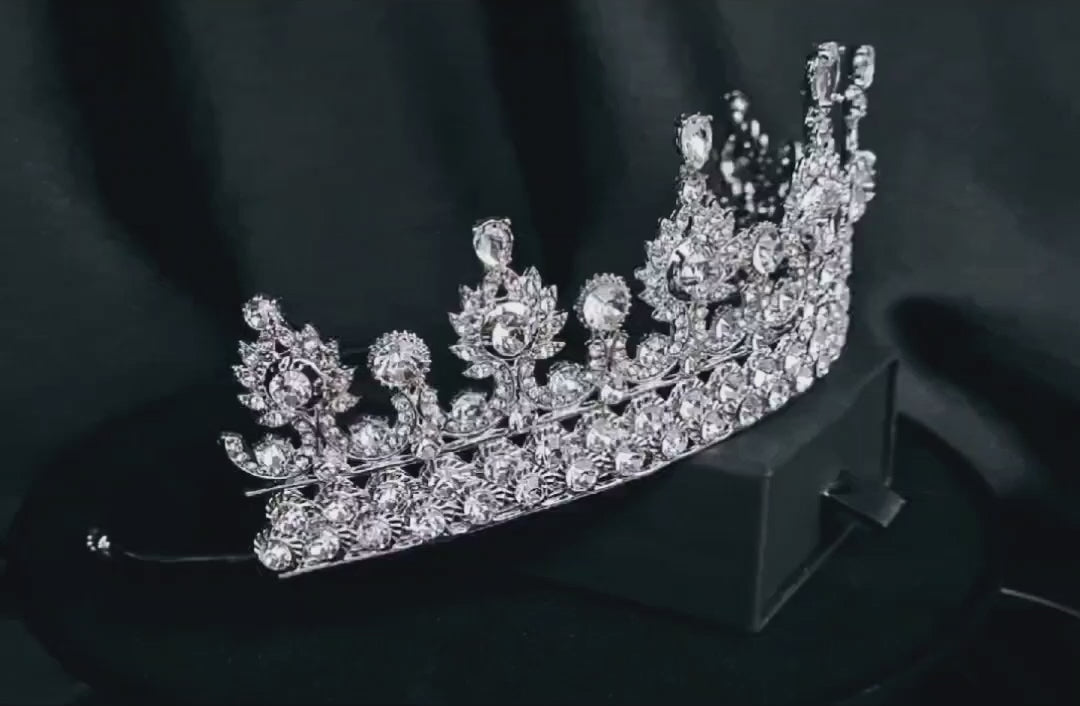 A close-up of a tiara with rhinestones on a black background. The tiara is made of silver and features a delicate design with cascading rhinestones. The rhinestones are clear and sparkling.