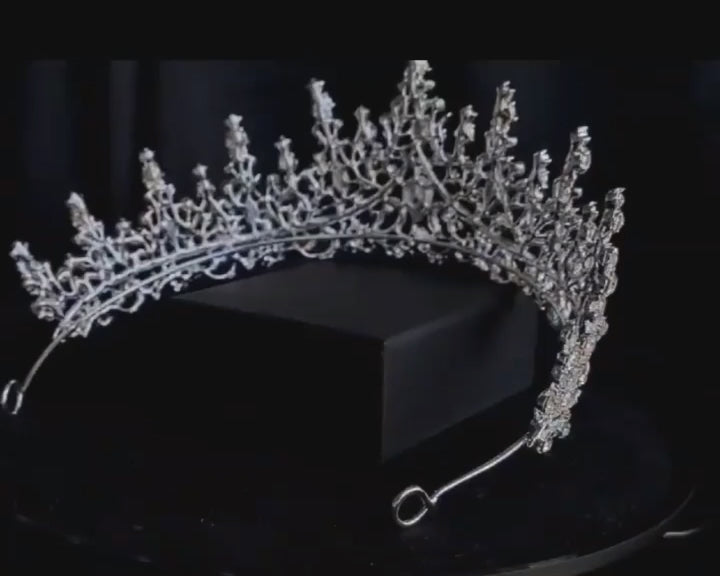 A close-up of a tiara with rhinestones on a black background. The tiara is made of silver and features a delicate design with cascading rhinestones. The rhinestones are clear and sparkling.