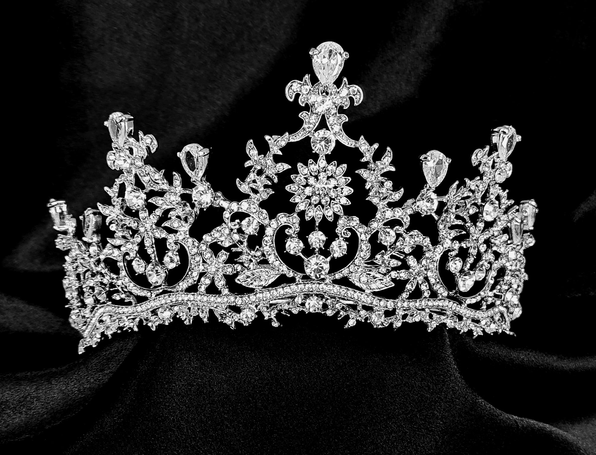 A silver tiara with rhinestones and flowers sitting on a black box. The tiara is made up of multiple curved bands, each of which is decorated with rhinestones and flowers. The tiara is sitting on top of a black box, which is partially obscuring the bottom of the tiara.