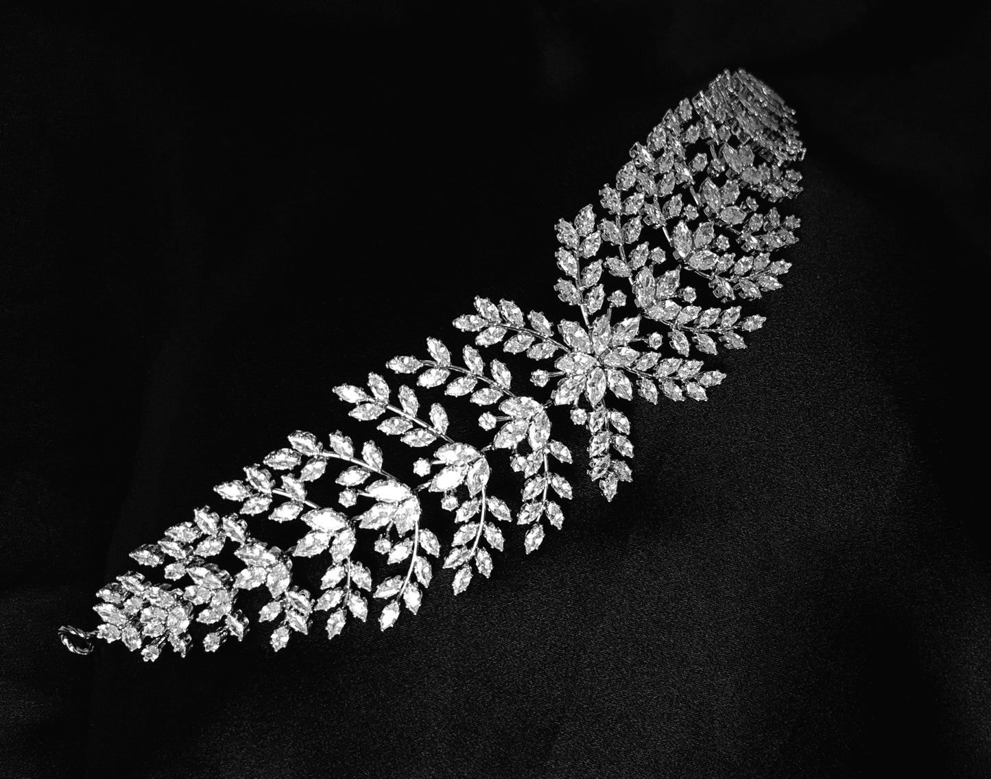 A wedding hair accessory made of metal and diamonds. The tiara has a delicate design with leaves and flowers. The diamonds are clear and sparkling. The background is black.