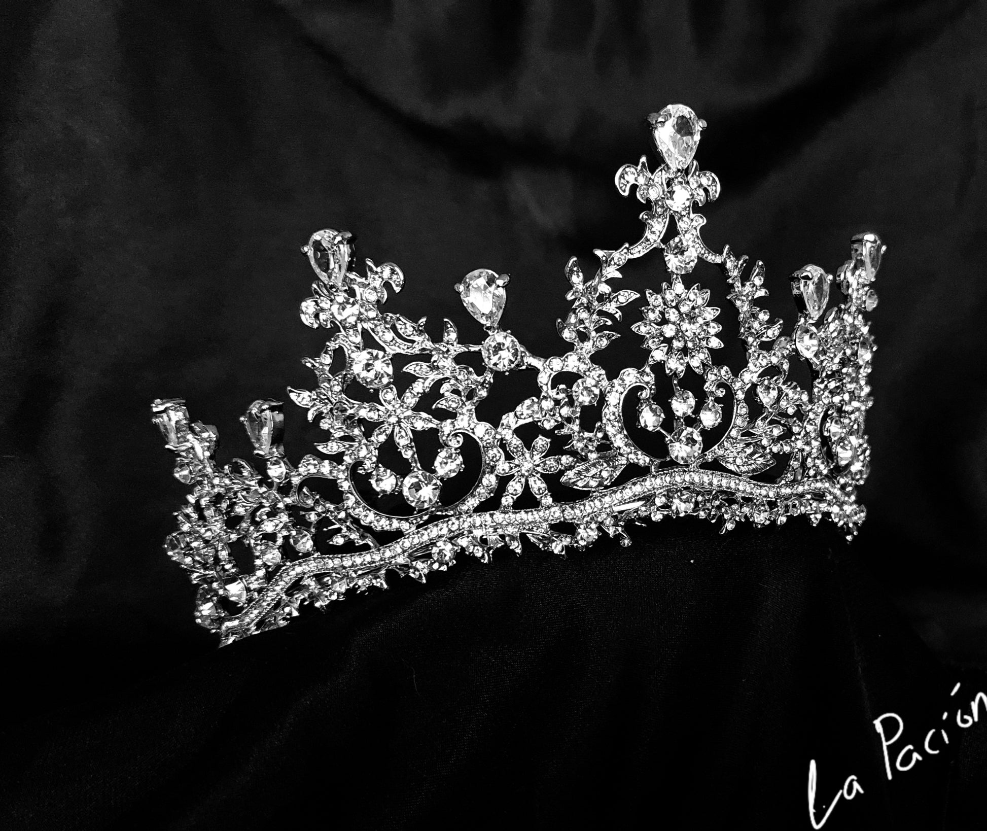 A silver tiara with rhinestones and flowers sitting on a black box. The tiara is made up of multiple curved bands, each of which is decorated with rhinestones and flowers. The tiara is sitting on top of a black box, which is partially obscuring the bottom of the tiara.