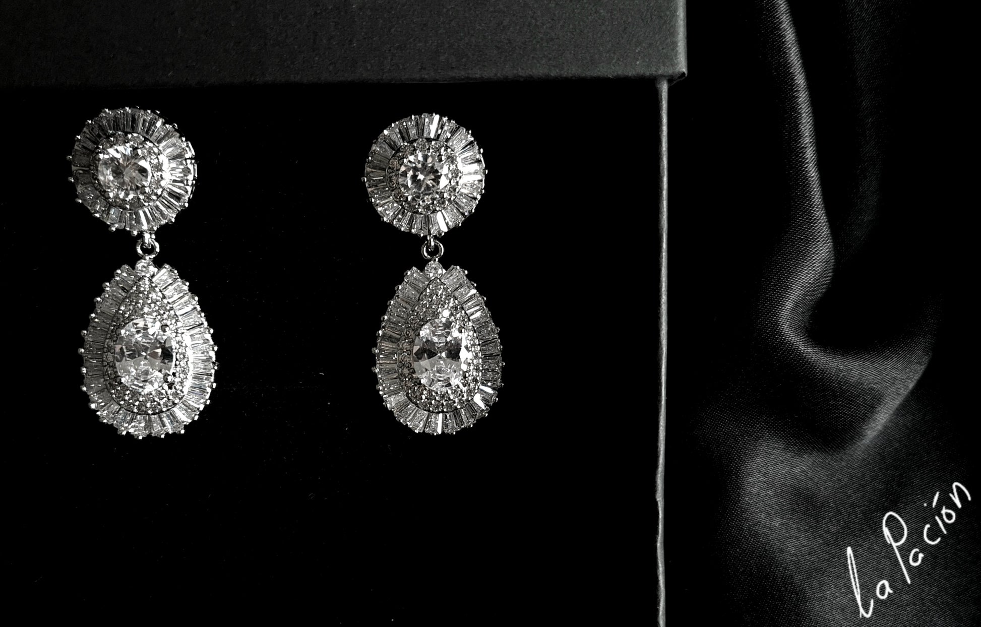A pair of dangle earrings made of cubic zirconia. The earrings are teardrop-shaped and have a simple, elegant design. The cubic zirconia stones are clear and sparkling. The earrings are hanging from a silver metal wire. The earrings are sitting on a black velvet surface.