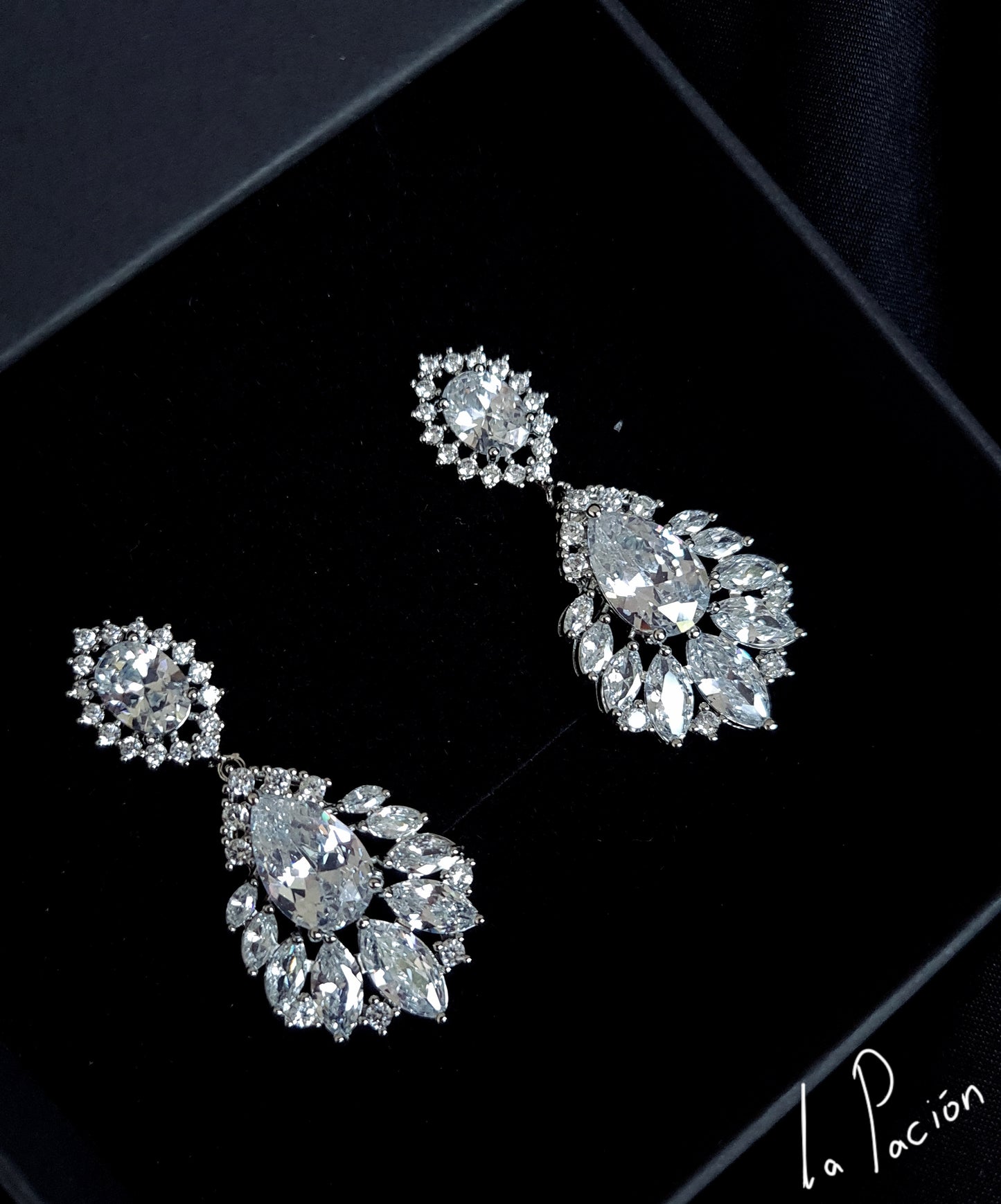 A pair of earrings with diamonds on a black background. The earrings are made of silver and have a delicate design. The diamonds are round and sparkling. The earrings are hanging from a silver chain.