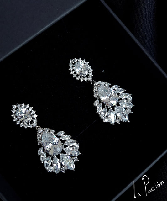 A pair of earrings with diamonds on a black background. The earrings are made of silver and have a delicate design. The diamonds are round and sparkling. The earrings are hanging from a silver chain.