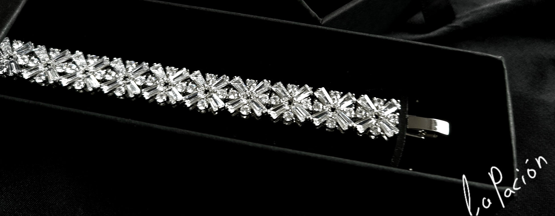 A bracelet with diamonds in a box on a black background. The bracelet is made of silver and has diamonds encrusted on it. The diamonds are sparkling and the bracelet is very shiny. The box is black.