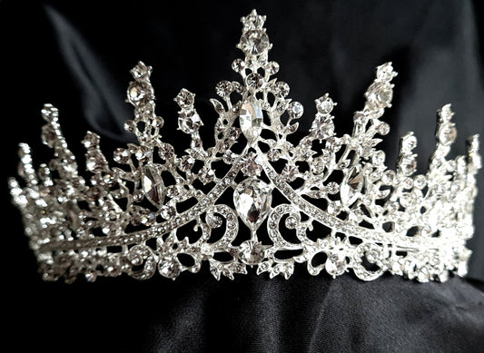 A close-up of a tiara with rhinestones on a black background. The tiara is made of silver and features a delicate design with cascading rhinestones. The rhinestones are clear and sparkling.