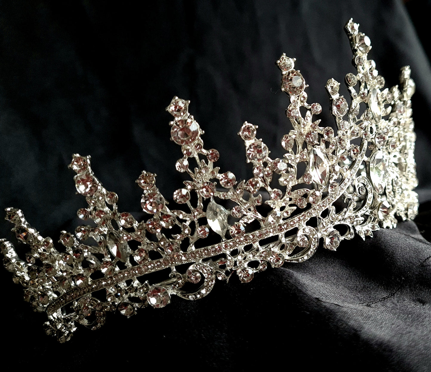 A close-up of a tiara with rhinestones on a black background. The tiara is made of silver and features a delicate design with cascading rhinestones. The rhinestones are clear and sparkling.