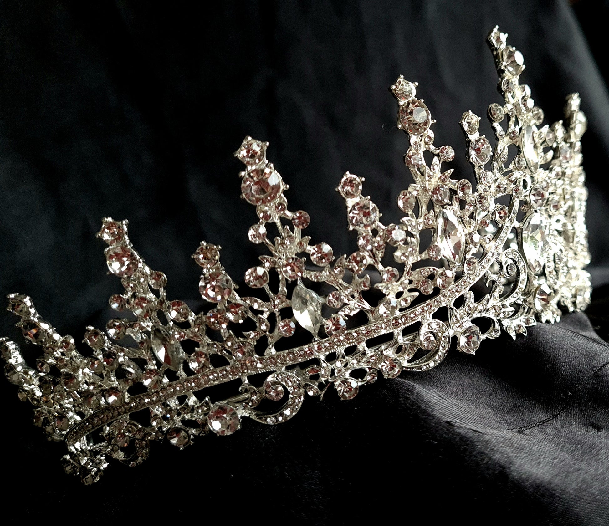 A close-up of a tiara with rhinestones on a black background. The tiara is made of silver and features a delicate design with cascading rhinestones. The rhinestones are clear and sparkling.