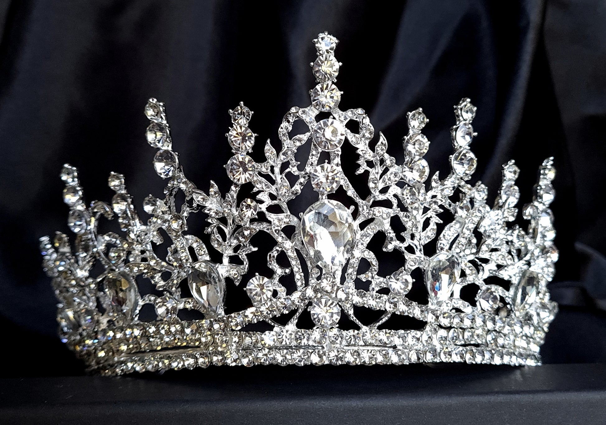 A close-up of a tiara with rhinestones on a black background. The tiara is made of silver and features a delicate design with cascading rhinestones. The rhinestones are clear and sparkling.