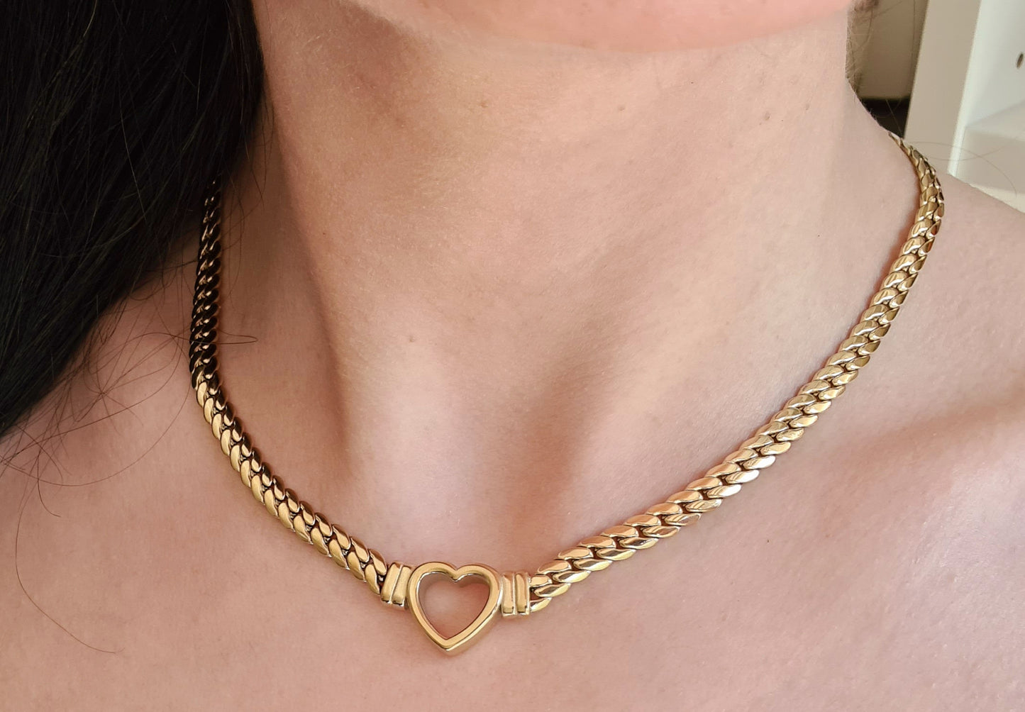 Woman wearing a Gold necklace with heart pendant, the necklace is made of gold and has a heart pendant in the center. The heart is gold and has a smooth surface.