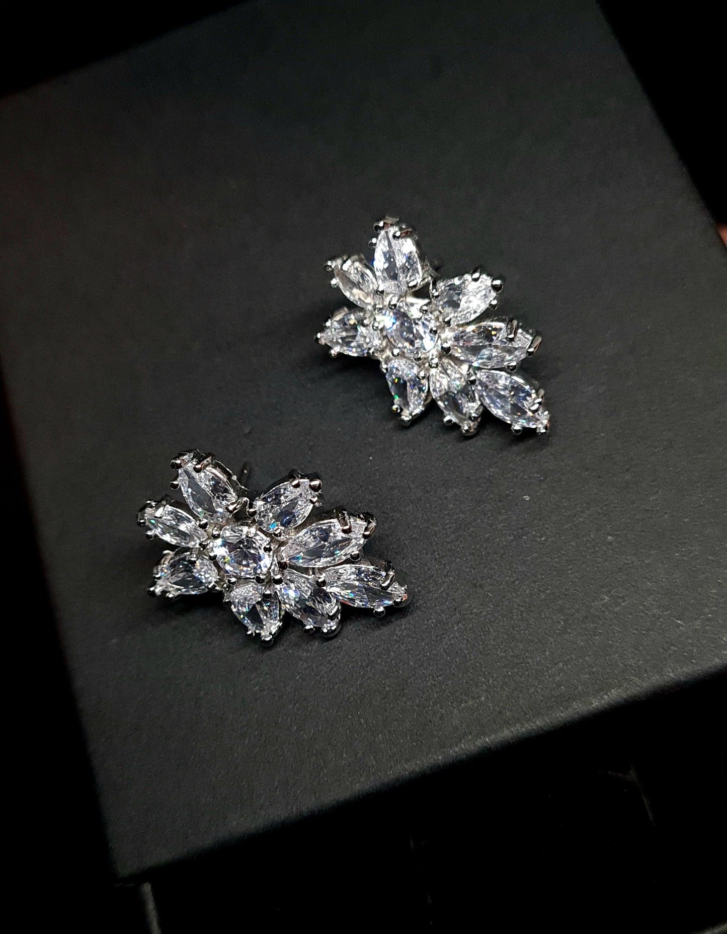 A pair of earrings in the shape of a flower, made from cubic zirconia stones. The earrings are sitting on a black surface. The flower is about 2.6 cm and is made up of petals that are clear and sparkling. The center of the flower is a large cubic zirconia stone.