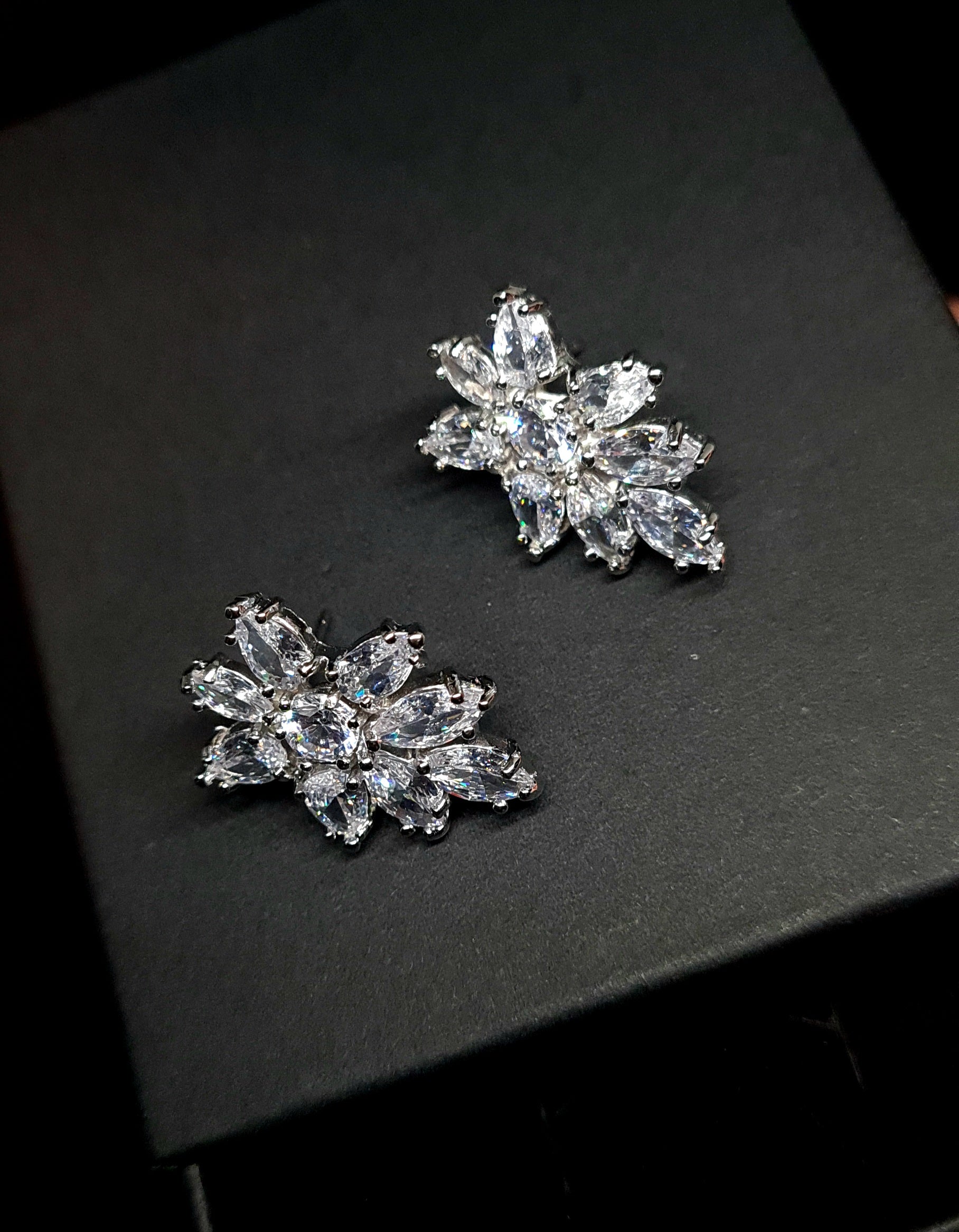 A pair of earrings in the shape of a flower, made from cubic zirconia stones. The earrings are sitting on a black surface. The flower is about 2.6 cm and is made up of petals that are clear and sparkling. The center of the flower is a large cubic zirconia stone.
