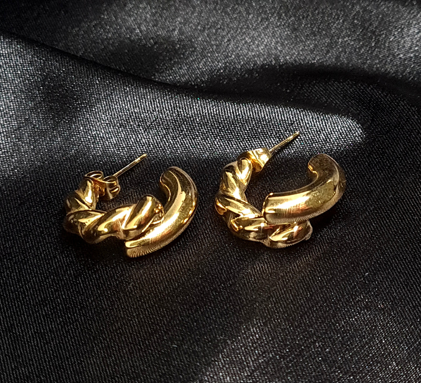 Gold hoop earrings sitting on black cloth. The earrings are a classic and elegant style that would be perfect for any occasion. The earrings are made of gold and have a hoop design. The hoops are large and round, and they sit on a black cloth.