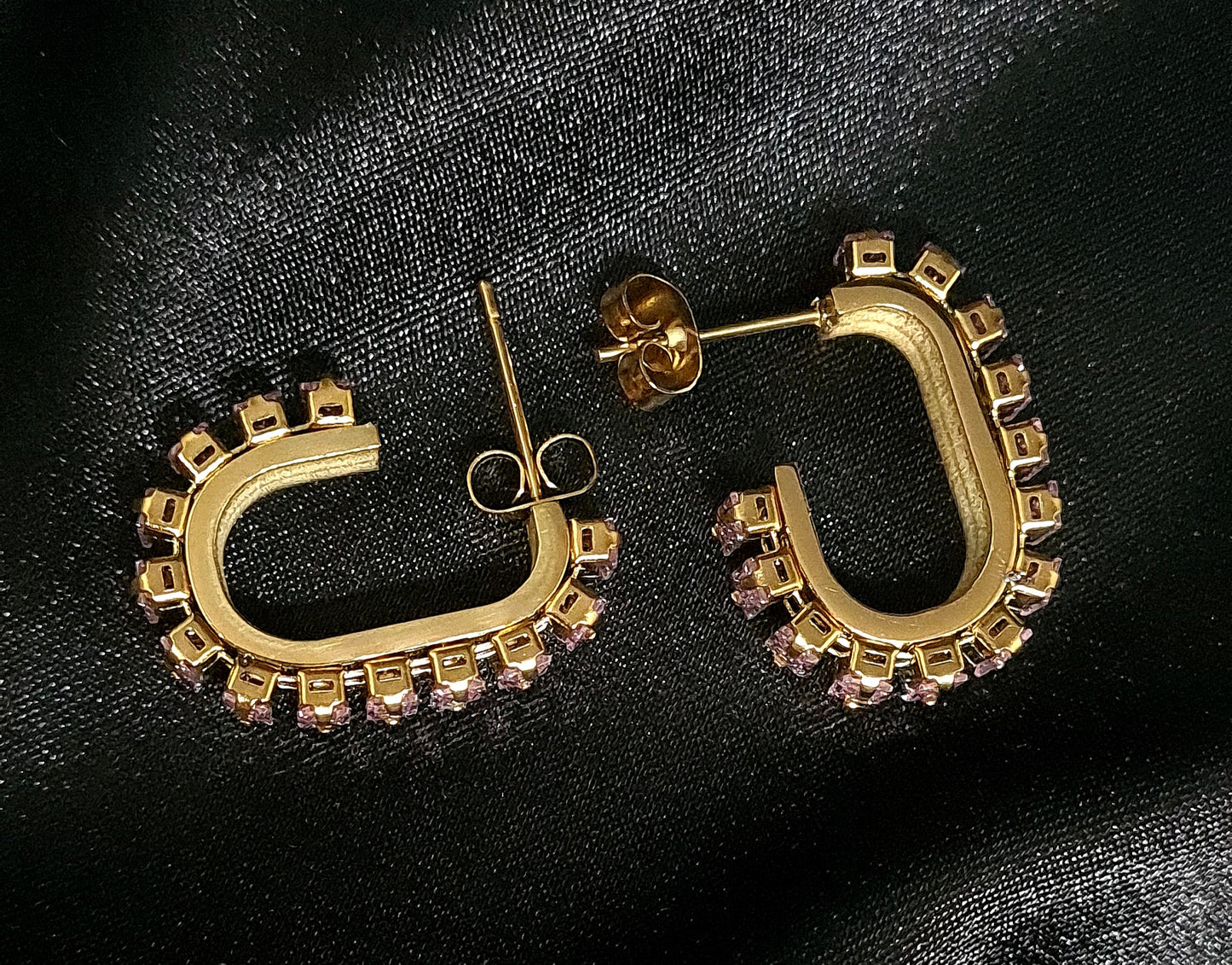 A pair of gold geometric design hoop earrings with pink cubic zirconia stones on a black cloth.